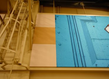 Blue image panel set within a tan wall system beside exposed structure.